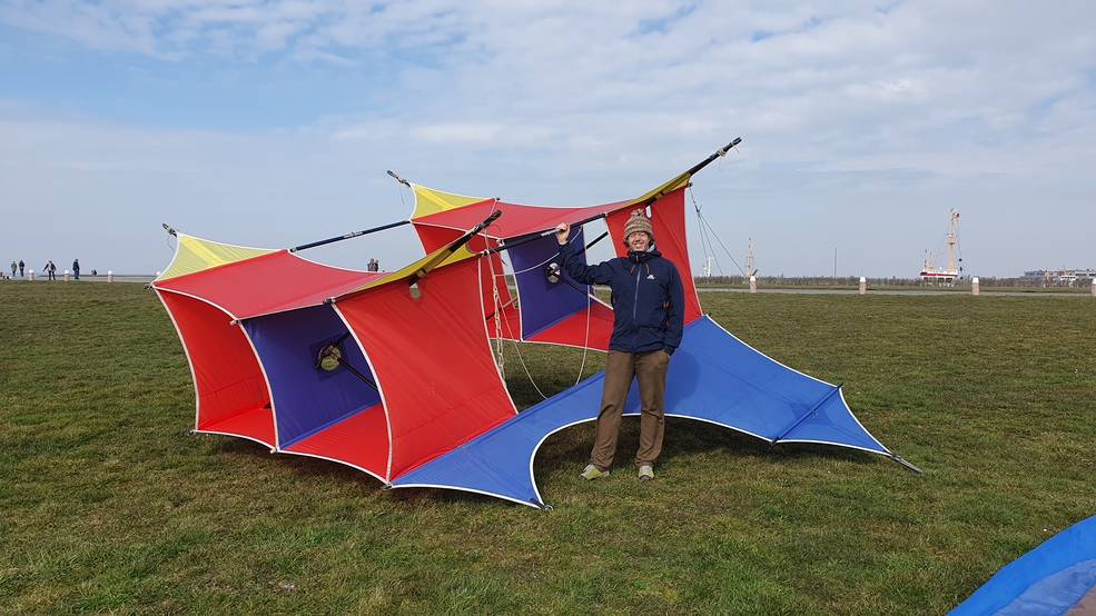 A large box-shaped kite with wings, colored red, blue, and yellow, sits on a large field of grass. A person wearing a blue coat, brown pants, and a brown hat stands next to the kite and smiles at the camera.