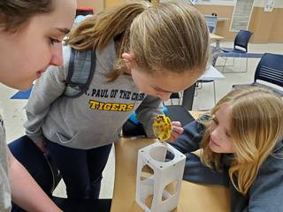 Three female students inspect their experiment. One holds a yellow, cone-shaped component with flat metal pieces adhering to its convex side.