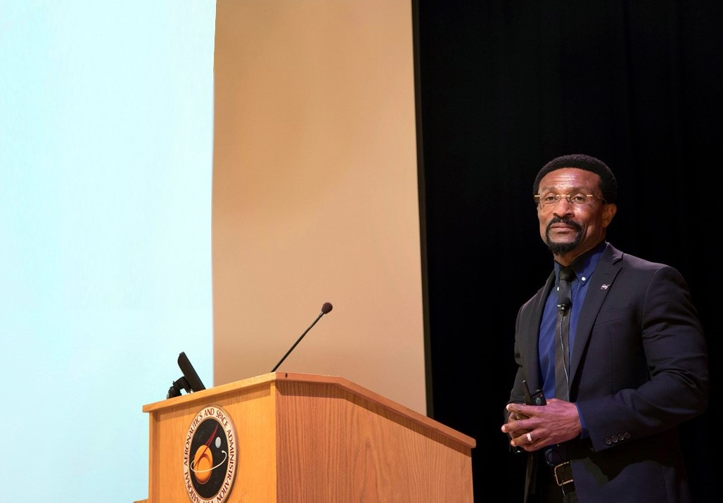 Moses Adoko, a man with short, curly black hair and beard, stands behind a lectern and looks out toward the left of the image, hands folded in front. He wears a black suit and tie with blue shirt, and stands in front of a black curtain and white screen.