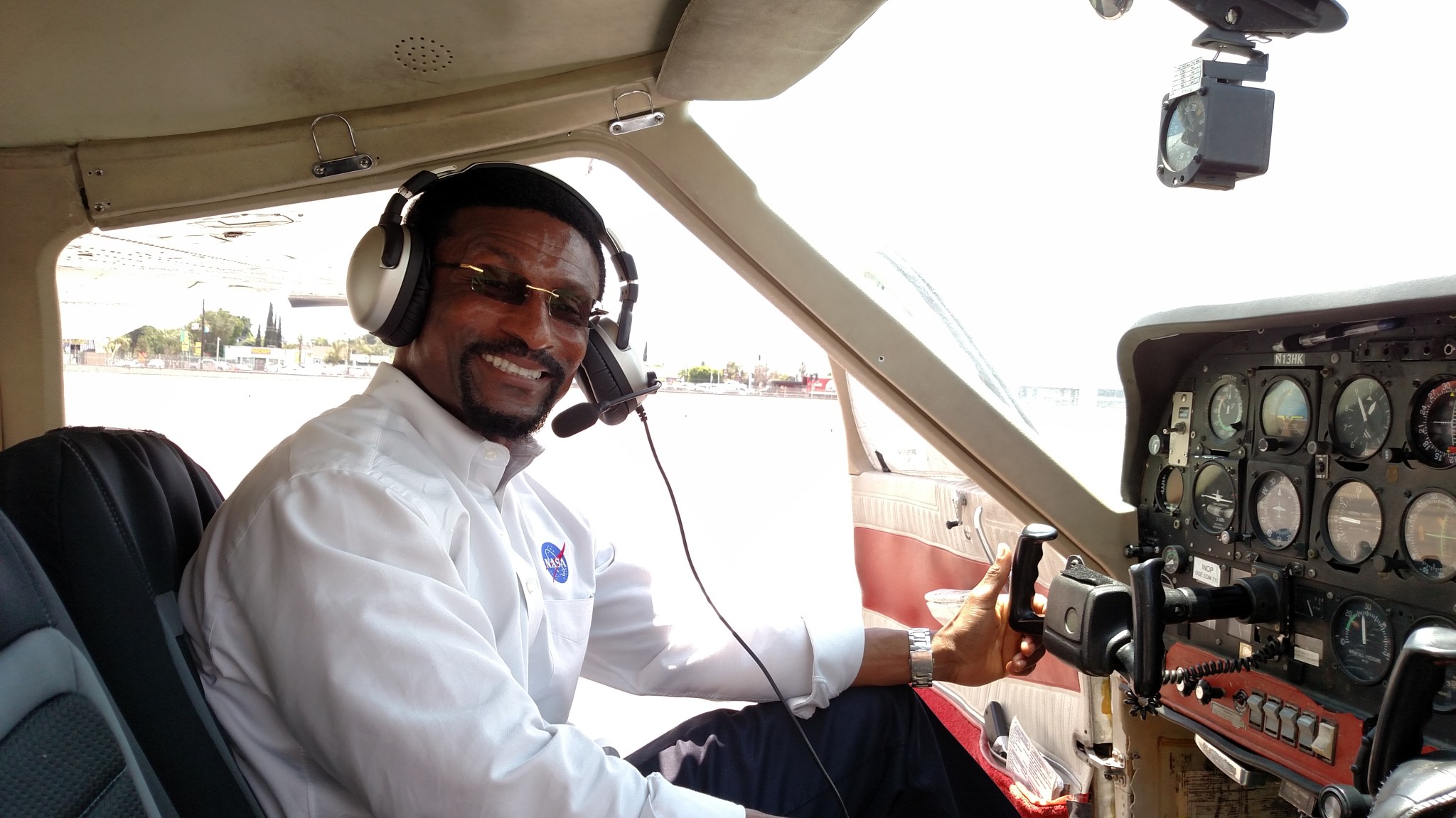 Moses Adoko, a man with short, curly black hair and beard, smiles and sits in a small airplane cockpit. He wears a headset, a white shirt with the NASA logo, and glasses. The plane door is open, showing outlines of trees and buildings.
