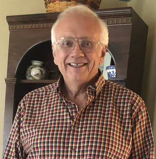 Bill Ochs, a man with white hair and glasses, smiles at the camera in a snapshot. He wears a red and brown checked shirt and stands in front of a bookcase filled with vases.