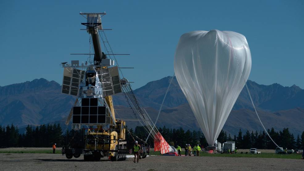 A scientific balloon is to the right, and appears as a plastic, upside down teardrop. A tube attached to the top of the balloon leads down to the ground. A crane to the left holds a large payload structure with many solar panels.