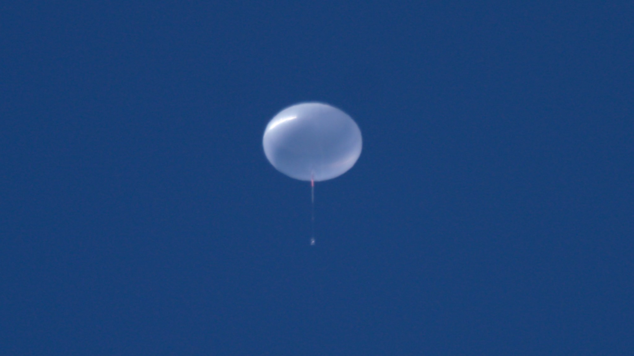A scientific balloon fully inflated floating in the sky. It appears plastic and clear against a clear blue sky.