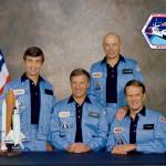 Four men in blue NASA outfits pose with model shuttle and U.S. flag