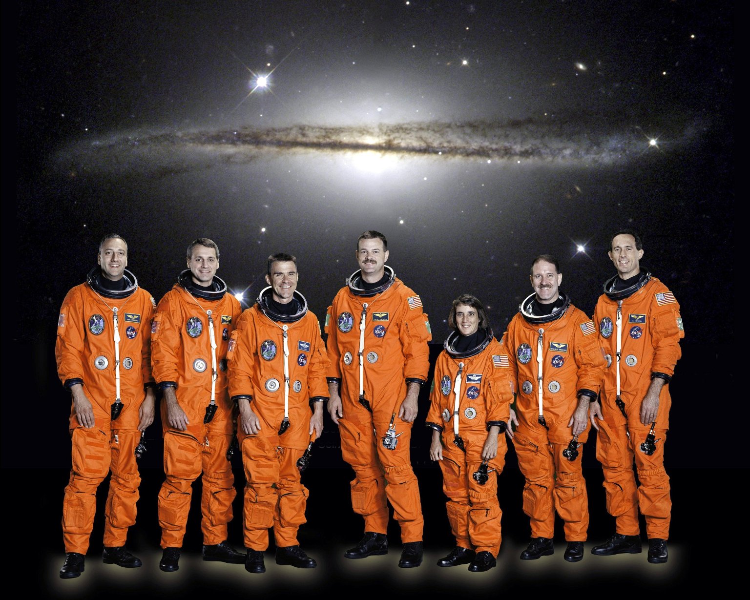 Six men and one woman in flight suits, no helmets, standing in front of a backdrop of a galaxy image.