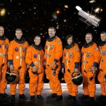 Six men and one woman in orange flight suits, some holding their helmets, standing in front of a space background with the Hubble Space Telescope in the image.