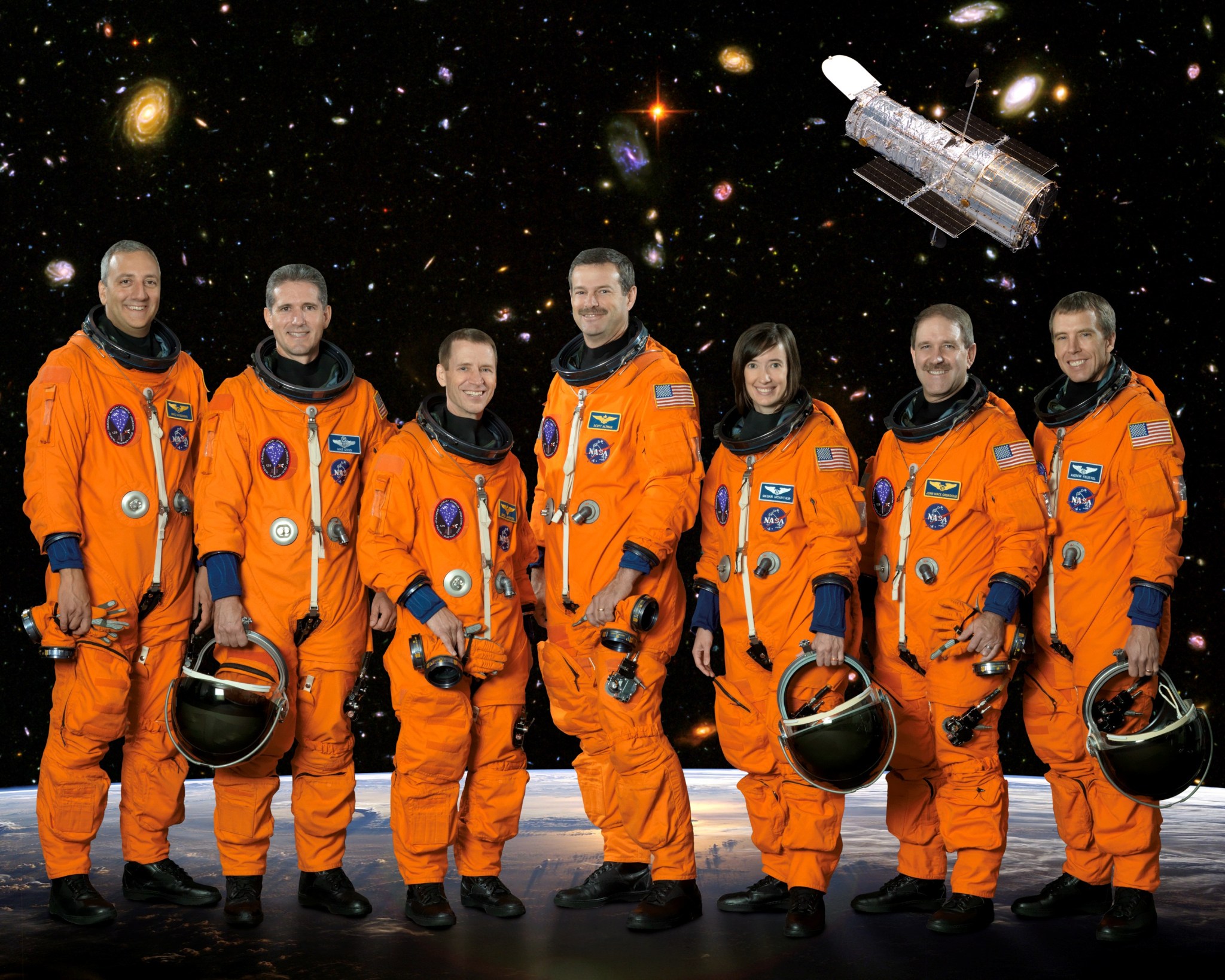 Six men and one woman in orange flight suits, some holding their helmets, standing in front of a space background with the Hubble Space Telescope in the image.