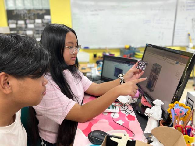 Two students at a table compare a piece of their experiment hardware to an image on a computer monitor. One student holds the hardware and points at the monitor.