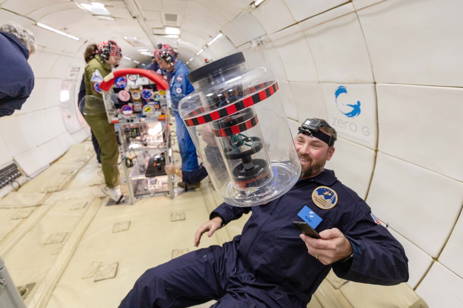 A device with black rings in a clear container floats in microgravity; a researcher with a head-mounted camera smiles at it. Background: Two researchers with surgical skull caps conduct experiments, their feet tucked under black straps to prevent floating.