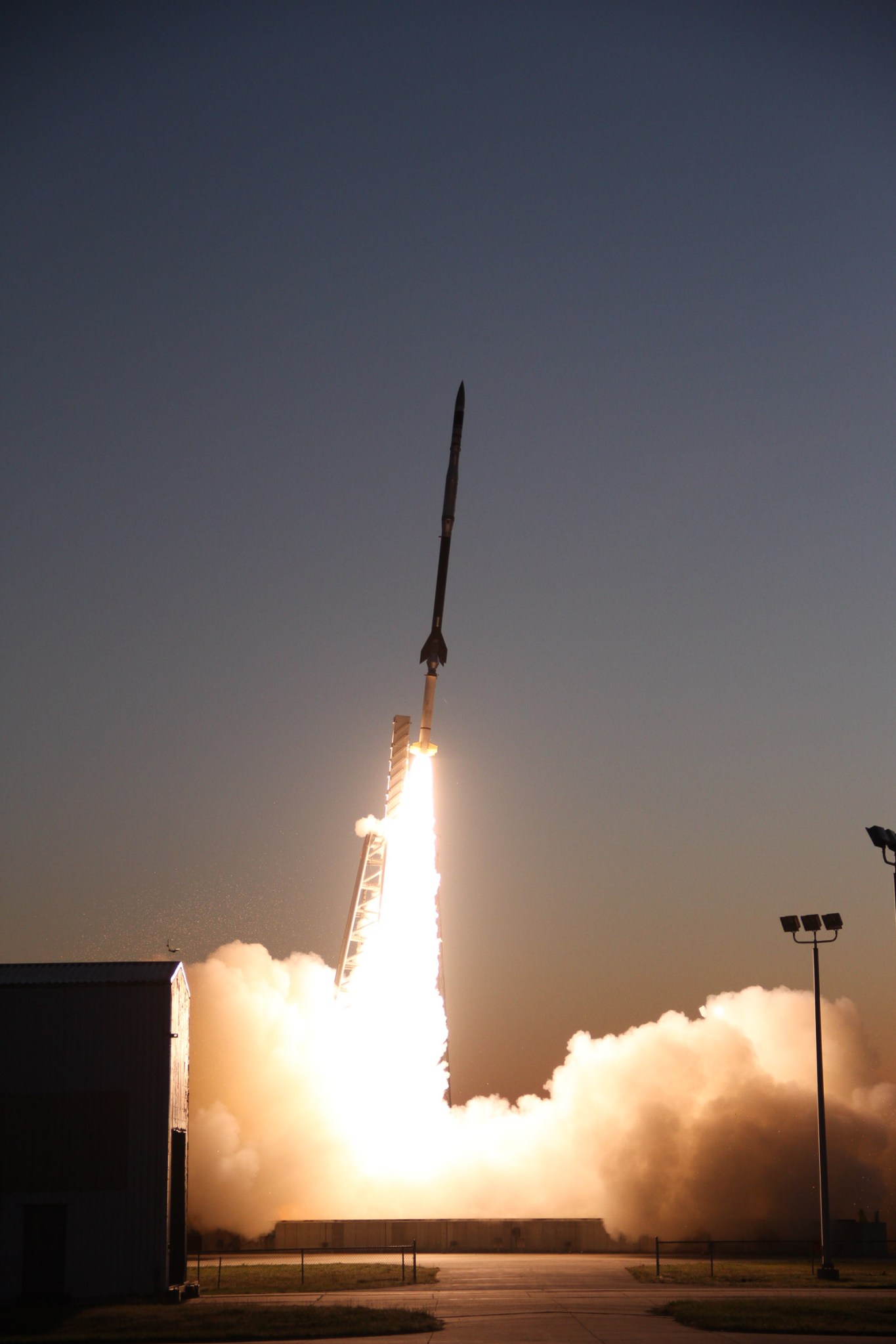 A sounding rocket seconds in mid-launch off the launch pad with a bright white plume the spreads out underneath the rocket when it hits the ground. The rocket is white on the bottom and darker up to the top.