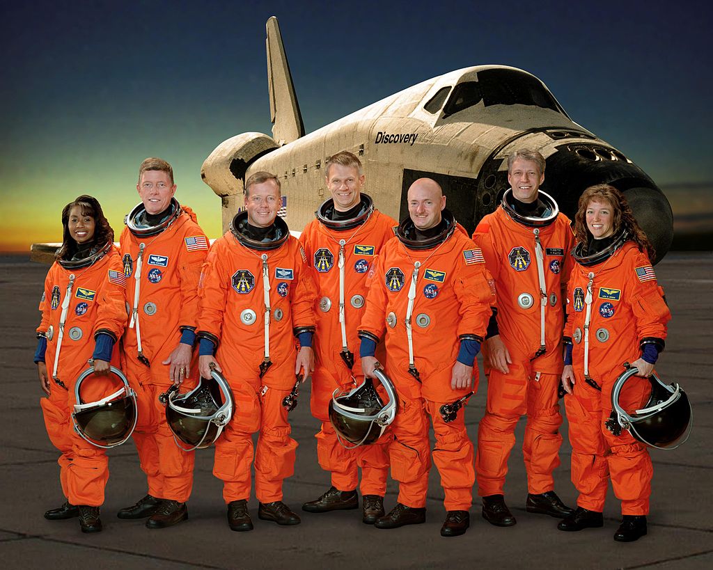 STS-121 crew in orange flight suits pose before the Discovery Space Shuttle
