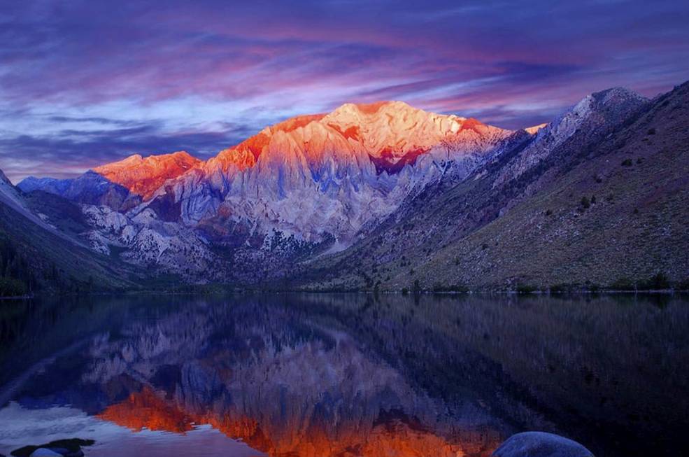 A colorful photo of a mountain and lake at sunset, its peak bright orange fading to deep purple near the water. The sky is streaked with blue and purple clouds.