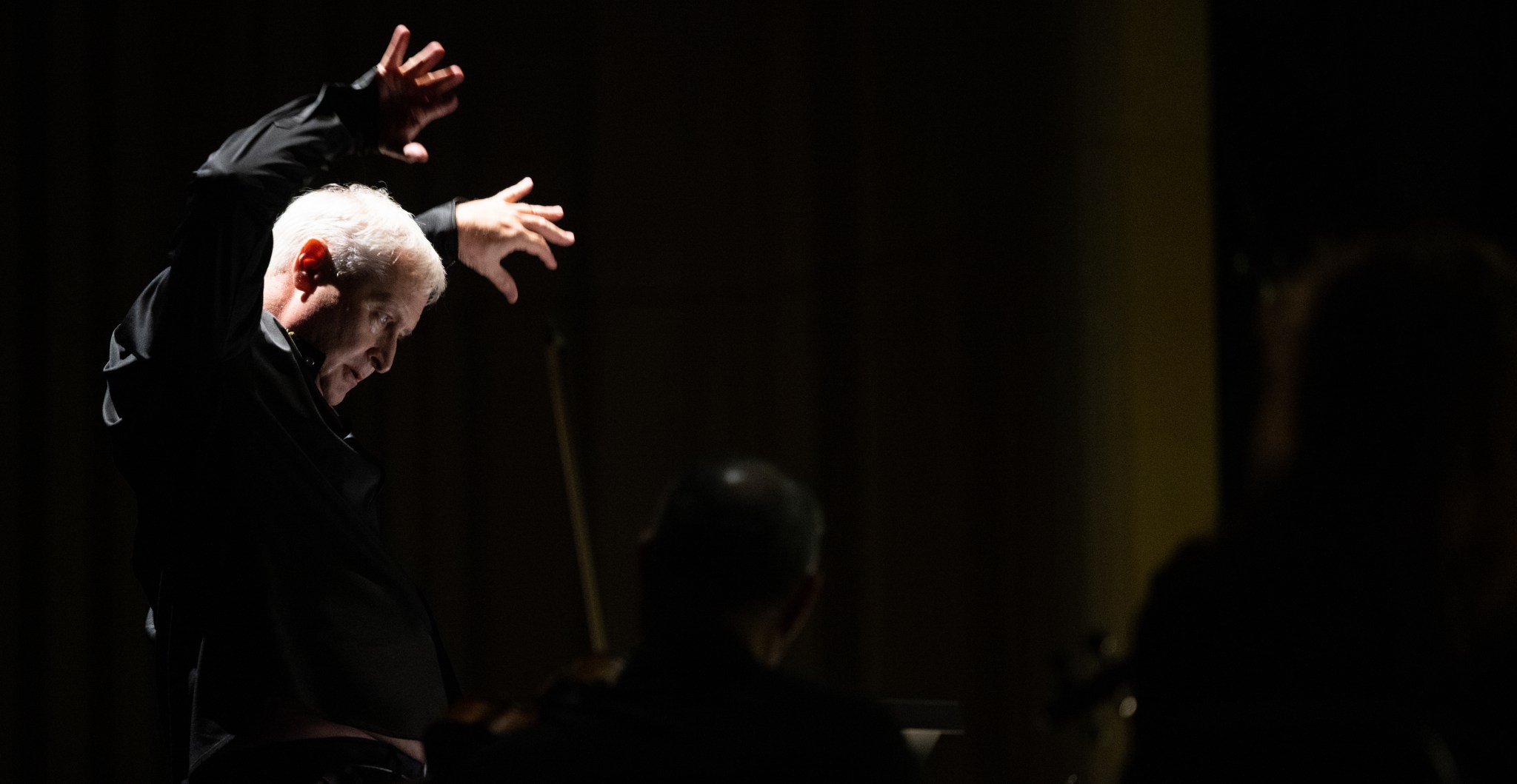Against a black background, a man with white hair waves his hands as he conducts an orchestra, off screen. He is looking downward toward a podium and his hands are blurred in motion, fingers spread wide.