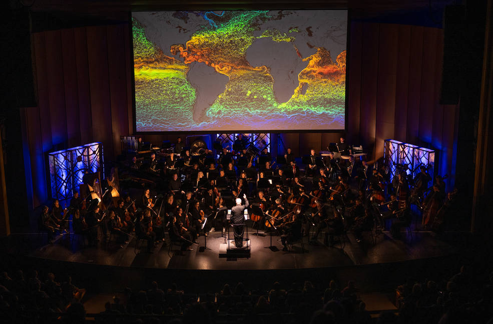 An illustration of Earth's ocean currents appears on a large screen behind an orchestra on a dark stage. Continents are gray and ocean swirls range from orange to blue. The orchestra is barely visible; a soft light illuminates the conductor.
