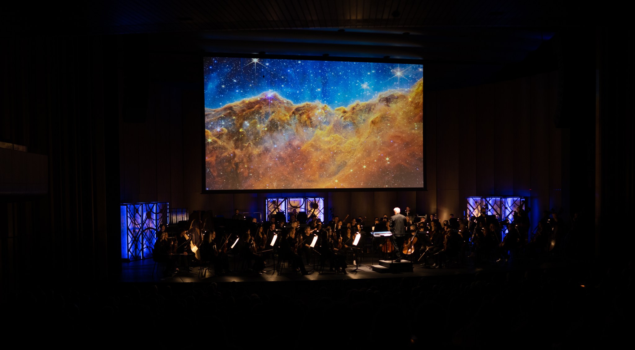 A darkened orchestra stage in a concert hall, with the James Webb Space Telescope image of the Carina Nebula projected on the screen. The theater appears mostly black, with the orchestra members faintly visible on stage and purple glowing panels lining the back of the stage. The image of the nebula looks like a bright orange cloud covering the bottom half of the screen, with jewel-tone blue and turquoise space visible behind. Bright stars sparkle throughout the image.