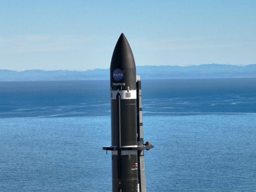 Close up of the top half of Rocket Lab's Electron rocket standing vertical at the launch pad with bright green grass and blue ocean waters in the distance. The black rocket's payload fairings have the NASA logo and TROPICS mission name on it. Credit: Rocket Lab