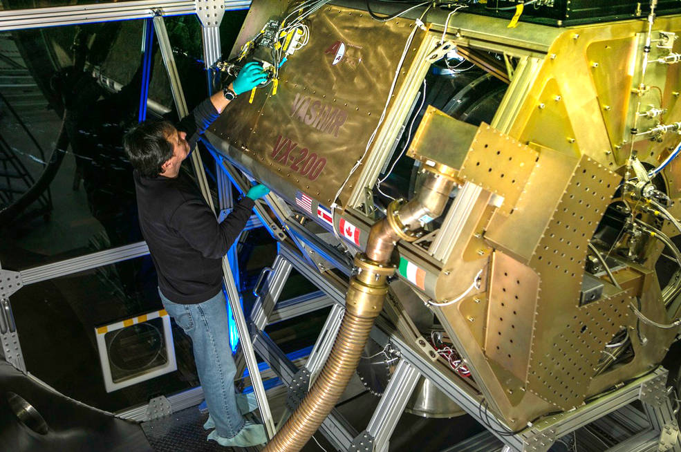 Man standing to the left of a medium-sized thermal vacuum chamber with his hand reaching up to adjust wires on the machine.