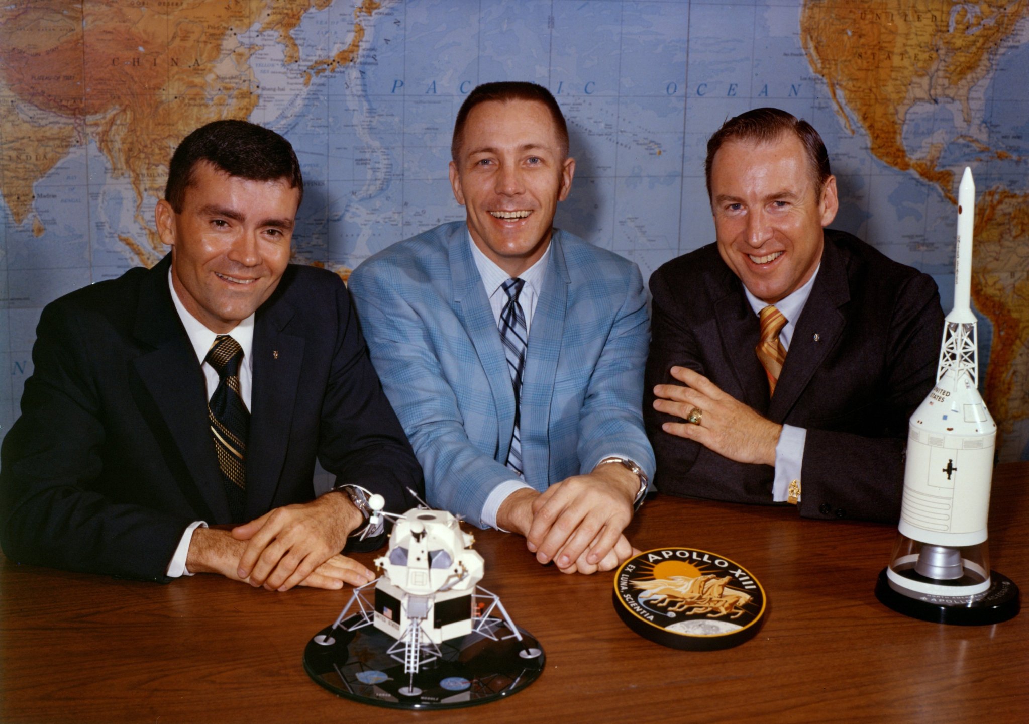 Three astronauts sit in suits at a table with the Apollo 13 patch and spacecraft replicas