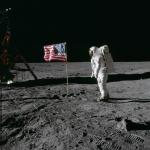 On the desolate pock-marked lunar surface, astronaut Edwin E. Aldrin Jr., wearing a white Extravehicular Mobility Unit spacesuit, stands to the right of the American flag planted in the soil. The flag is unfurled and waving to the left, with Aldrin facing it in the image and seen from a side view.