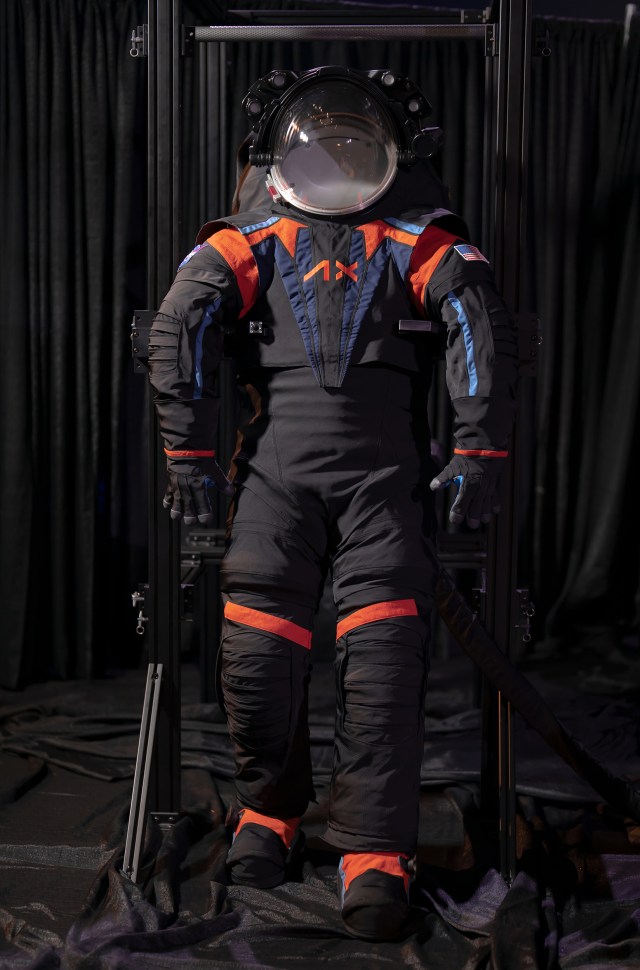 A full-body mock-up of Axiom's next generation spacesuit is shown, made of black material and orange accents.