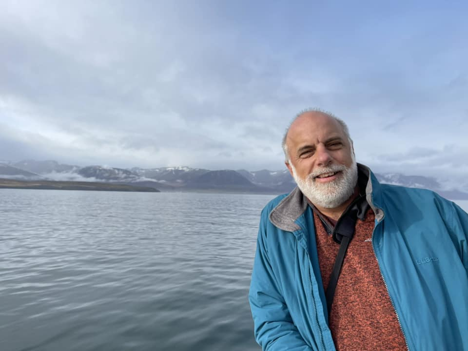 Randy Koster, a man with a white beard, smiles and leans into the picture in front of a background of gray-blue water, mountains, and clouds. He wears a burnt orange sweater and blue jacket.