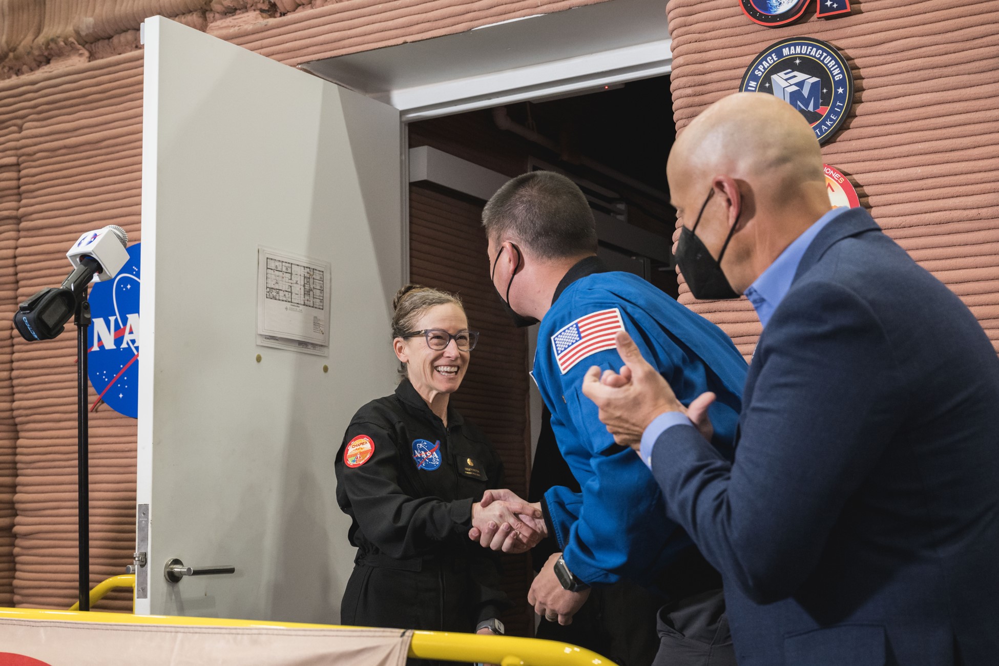 A woman in a black jumpsuit shakes the hand of a man in a blue jack. A man in a black suit stands and claps close by.