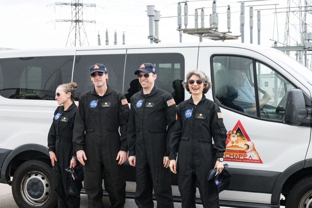 Four people in black jumpsuits stand outside of a white van