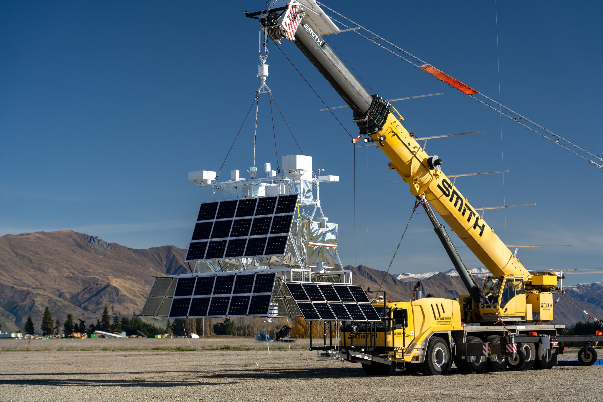 The EUSO-2 payload hangs from a yellow crane vehicle. The payload main body is mostly enclosed silver plastic wrapped wraps with multiple bars across the top with various instrumentation.