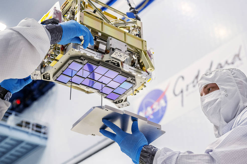 An iridescent purplish-blue array of squares attached to a chunky, rectangular piece of metal hardware is seen from below. Gloved hands guide the hardware while a man in a white suit and gloves attaches a silver covering to the array.