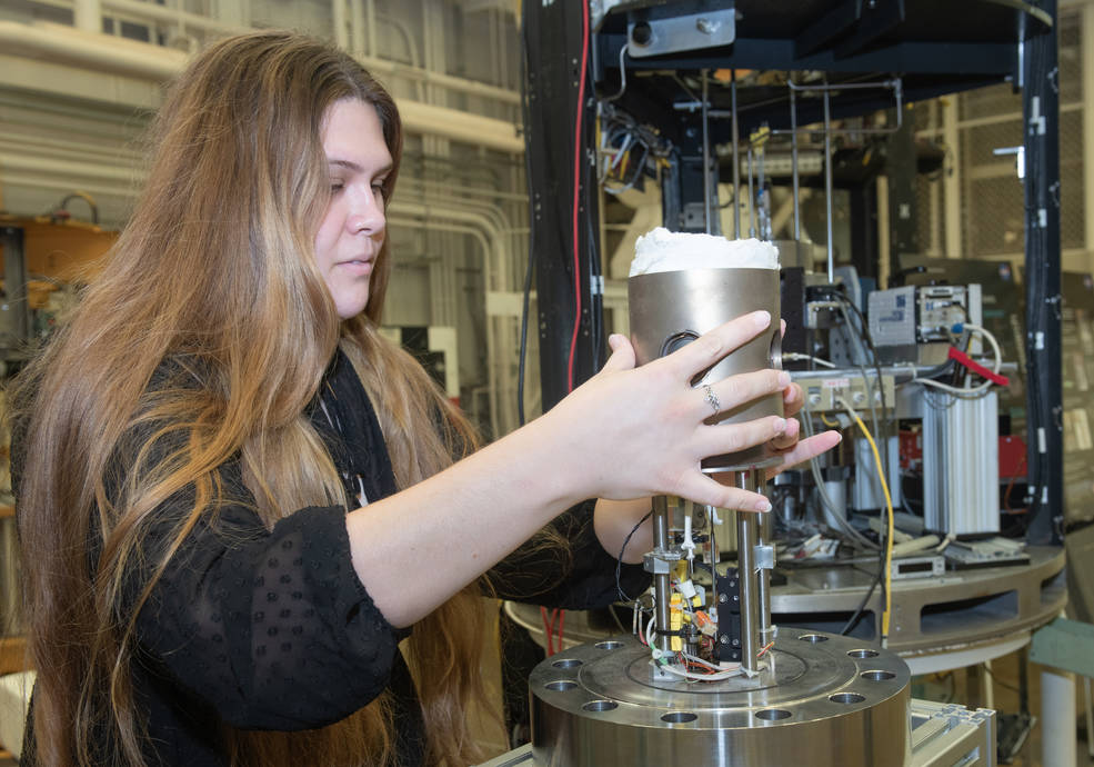 A female student with long brown hair and wearing a black top holds a large tube over to a smaller circular device with wires of all colors. In the background there is a large testing chamber with various sizes and colors of containers along with wires.