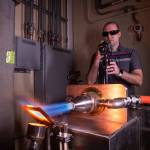 A man working in an industrial laboratory wearing sunglasses points a camera at a silver exhaust port emitting a superhot blue flame that is striking a material causing it to glow bright orange.