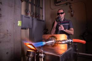 A man working in an industrial laboratory wearing sunglasses points a camera at a silver exhaust port emitting a superhot blue flame that is striking a material causing it to glow bright orange.