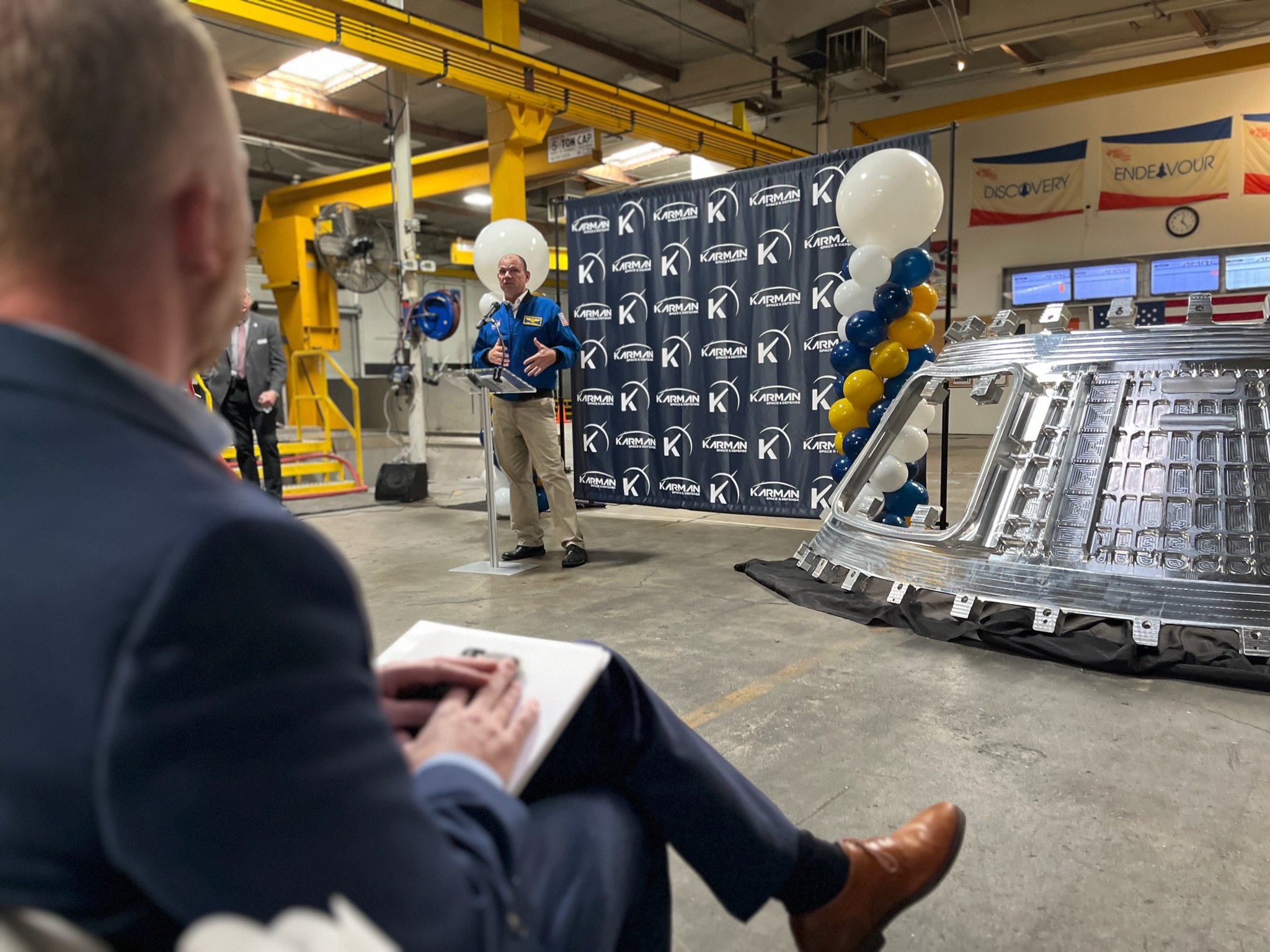 Retired NASA astronaut Tony Antonelli thanks employees at AMRO, Karman Space and Defense in South El Monte, California as a man in a suit sits with his legs crossed in the foreground.