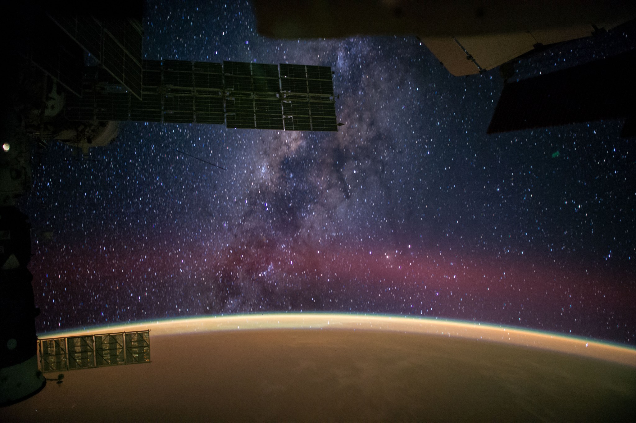 A panorama of the night sky and the Milky Way. There is a portion of the Earth visible showing the Sahara Desert golden sand with the purple and black image of space with thousands of white stars.