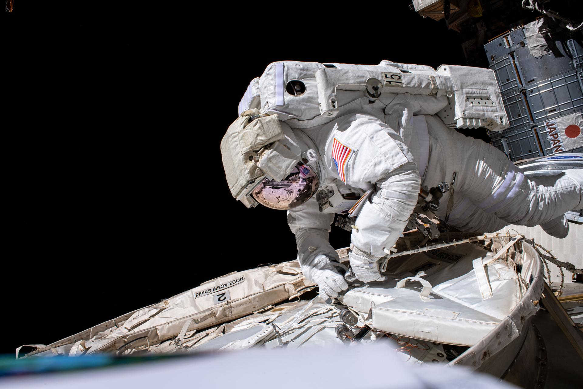 Against the black backdrop of the vacuum of space, NASA astronaut Chris Cassidy, entirely shielded by a spacesuit, works on maintenance outside the International Space Station. Using a wrench-type tool in his hands, he makes adjustments to a white outer layer of the orbiting laboratory.