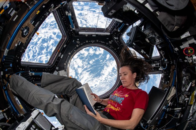 female astronaut reading a children's book while floating near the cupola of the space station with Earth on the backdrop