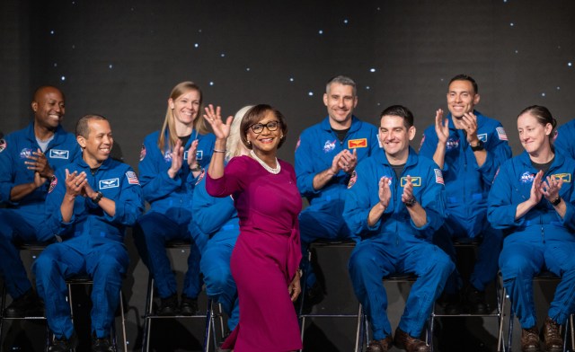 Center Director Vanessa Wyche in a burgundy dress waves to an audience with clapping astronauts sit behind her in their blue suits
