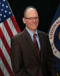 Mike Green wearing a suit standing in front of an American flag and a NASA flag.