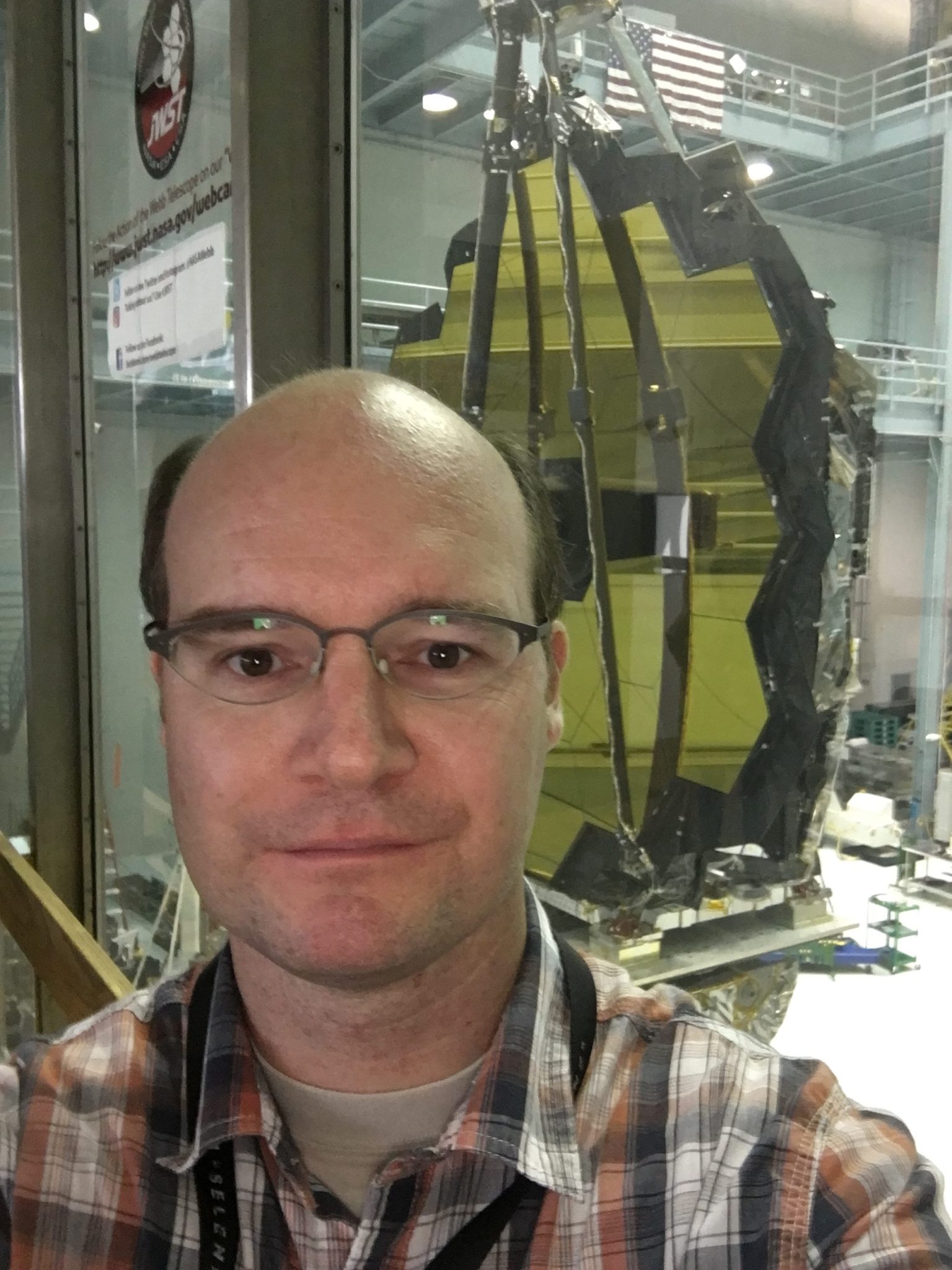 A man smiles slightly in a selfie in front of the James Webb Space Telescope's distinctive gold hexagonal mirror. He has dark hair above his hears and wears glasses an an orange, black, and white plaid shirt.