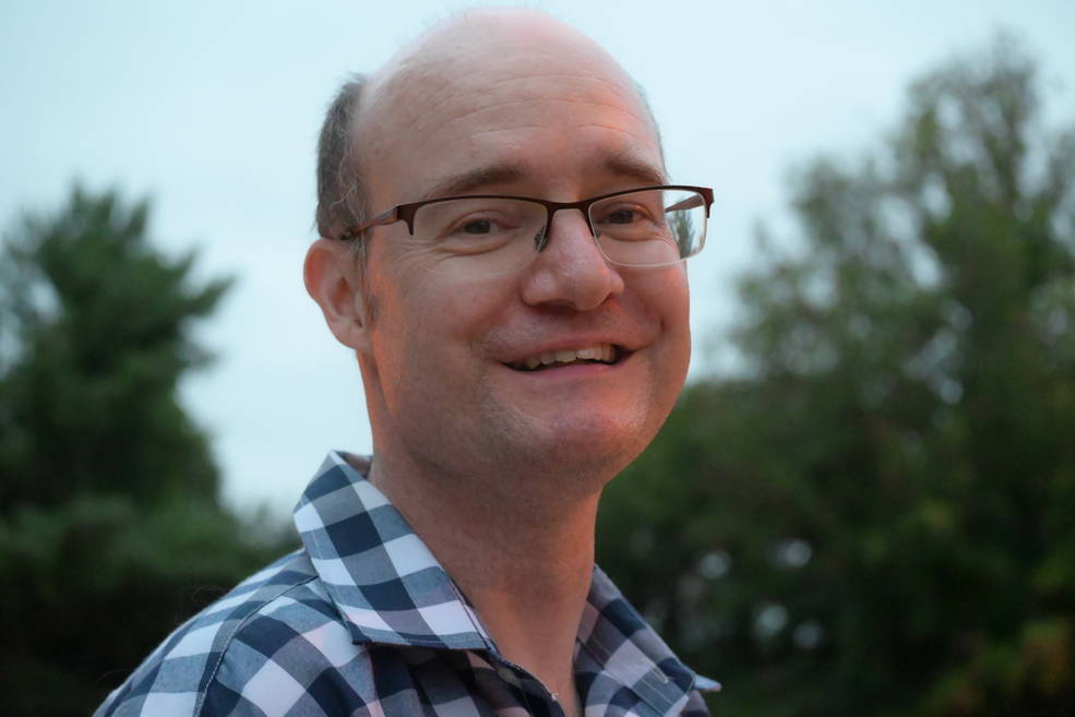 A man smiles in an artistic portrait. He has dark gray hair above his ears and wears glasses and a blue and white checked shirt. The light is low and bluish; trees are visible behind him, and an orange light reflects on his face.