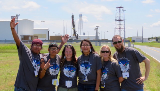 A group is six students wearing the same t-shirt the says "RockOn!" pose excitedly in front of sounding rocket standing vertical on the launch pad behind them.