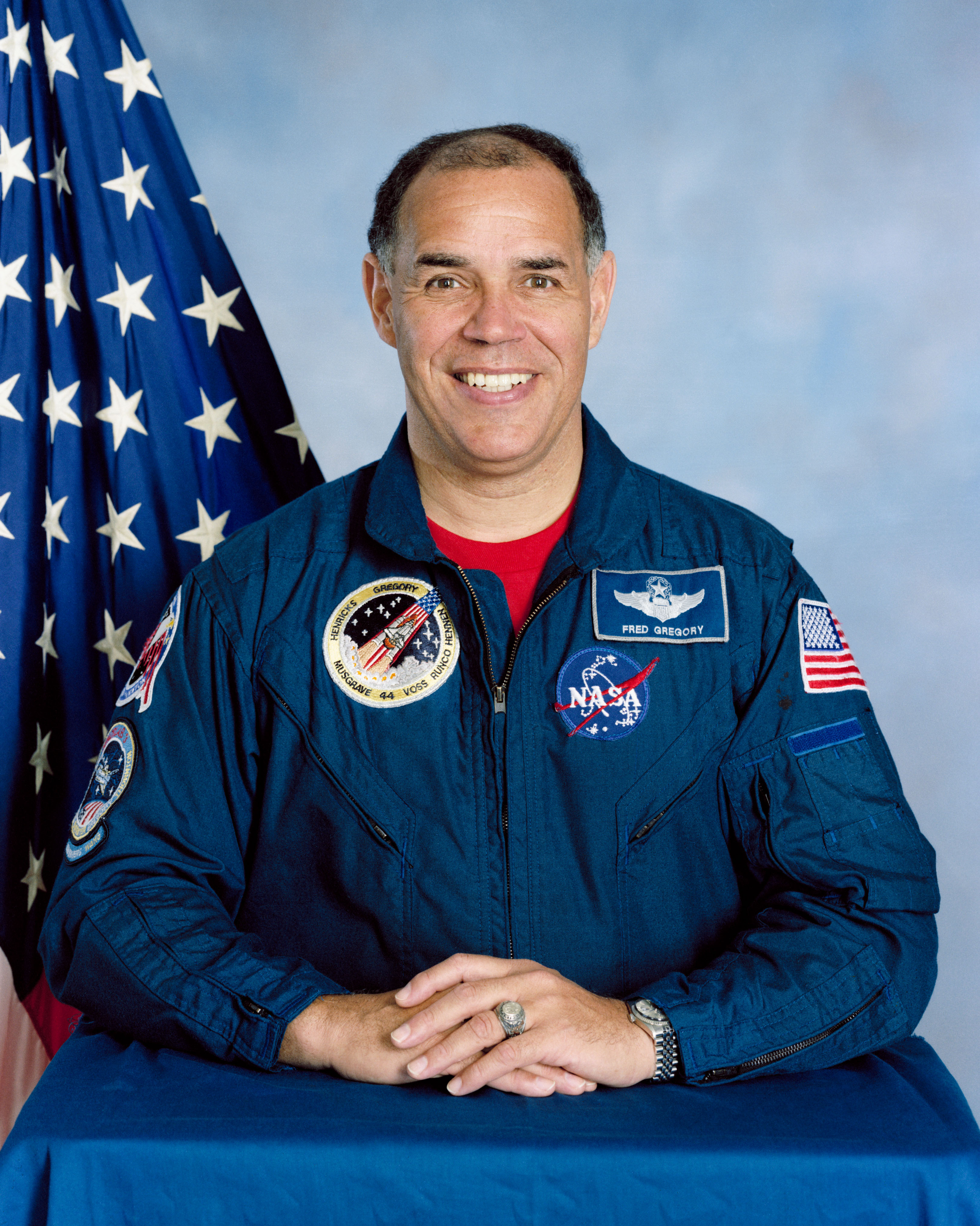 Official portrait of Frederick D. Gregory, a United States Air Force Colonel, wearing blue flight suit, with flag backdrop.