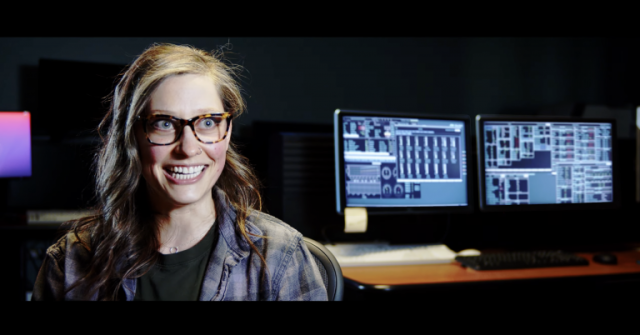 A young woman with blonde hair and glasses smiles at the camera with computer screens in the background.