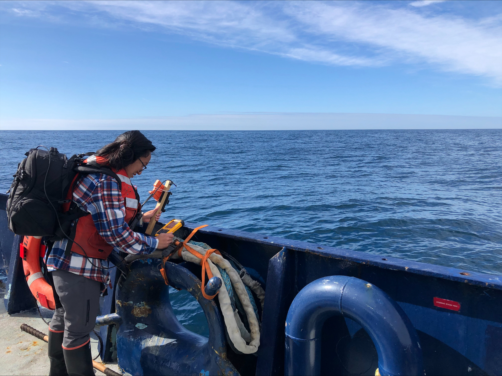 A field researcher stands at the edge of a boat overlooking the edge toward the ocean surface. She wears a backpack full of gear and holds an instrument facing the ocean.