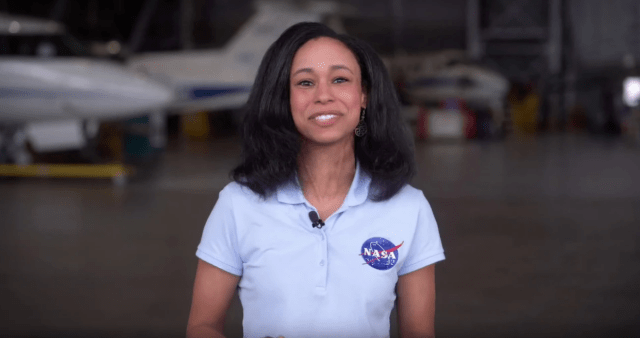 Small Steps Giant Leap's host on the Sonic Booms and the X-59. She is wearing a blue shirt with a NASA meatball on the right of her shirt. She is standing inside a hangar.