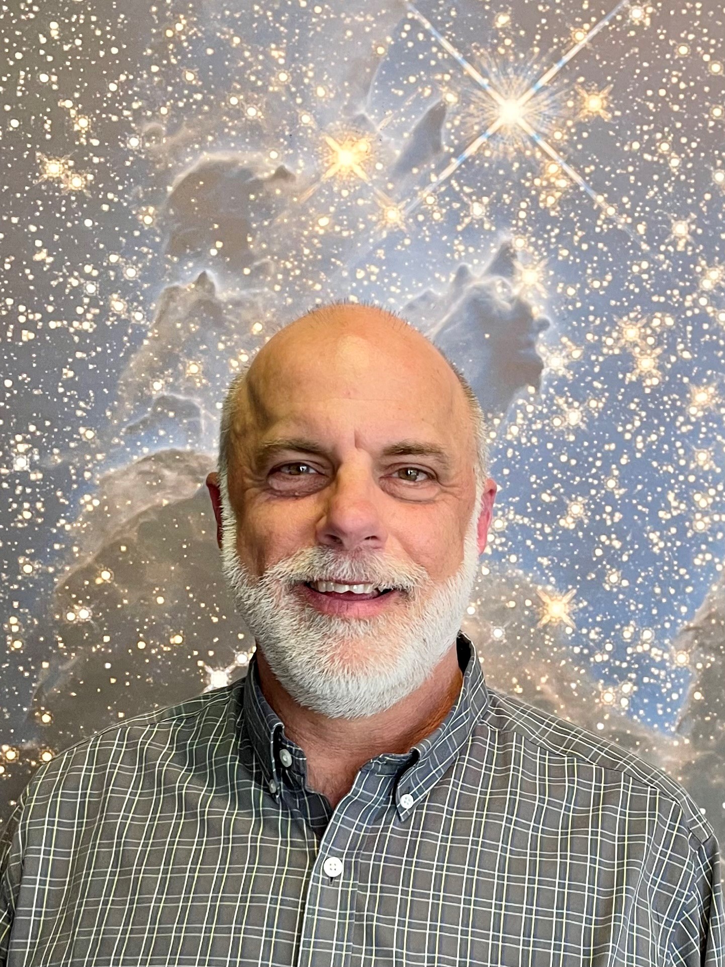 Randy Koster, a man with a white beard, smiles at the camera in a portrait. He wears a green checked shirt and stands in front of a faded galaxy background.
