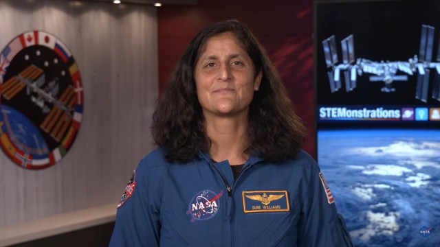 Astronaut Suni Williams, wearing a blue flight suit with a NASA patch and her name patch visible on the front, stands in a room with the International Space Station logo to her left and a visual containing an image of the International Space Station flying above Earth with the word "STEMonstrations" below it to her right.