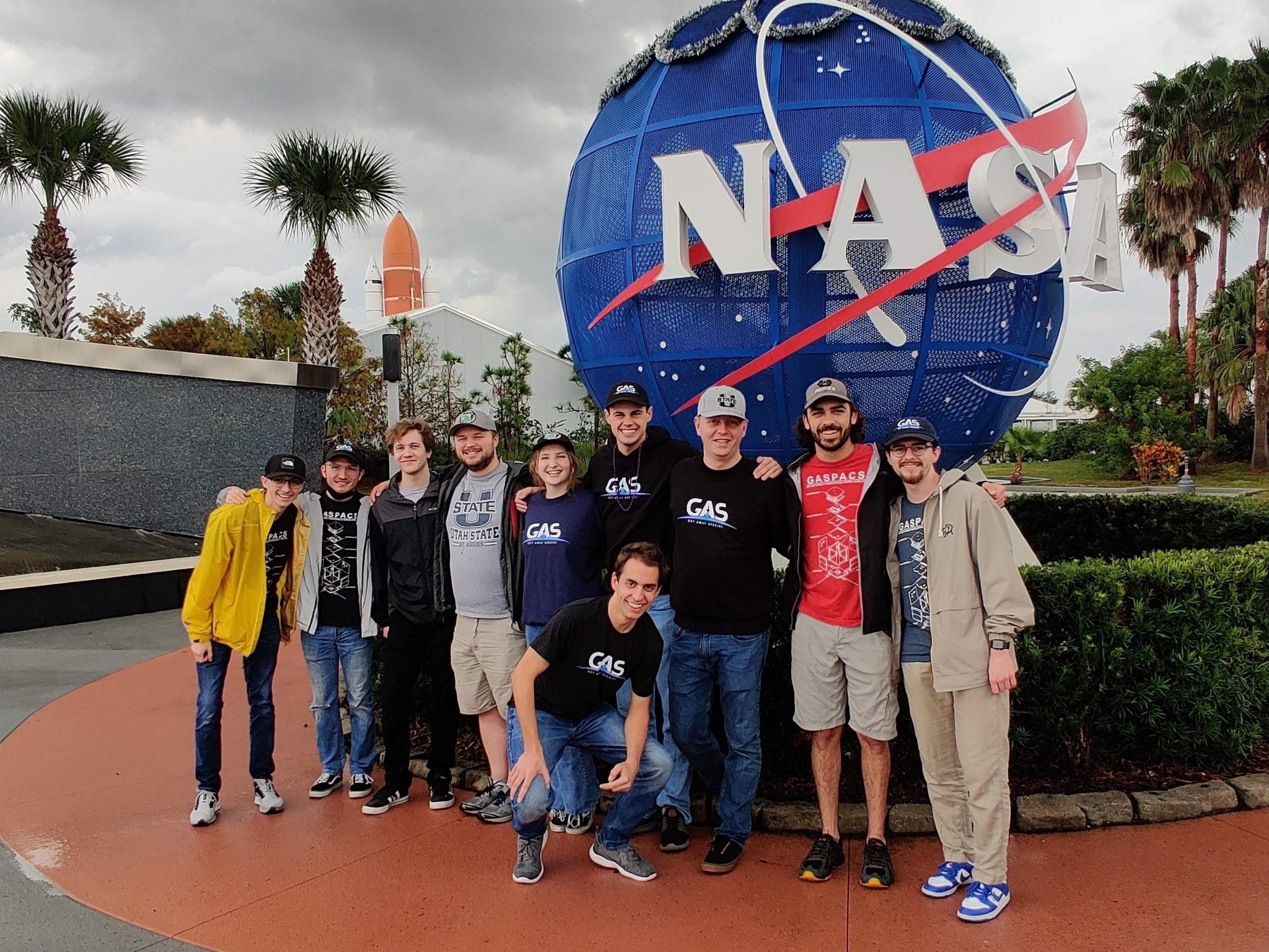 A group of students stand in front of a NASA sign with various bushes and plants around, in the very background is the top of the solid booster rocket on the launch pad.