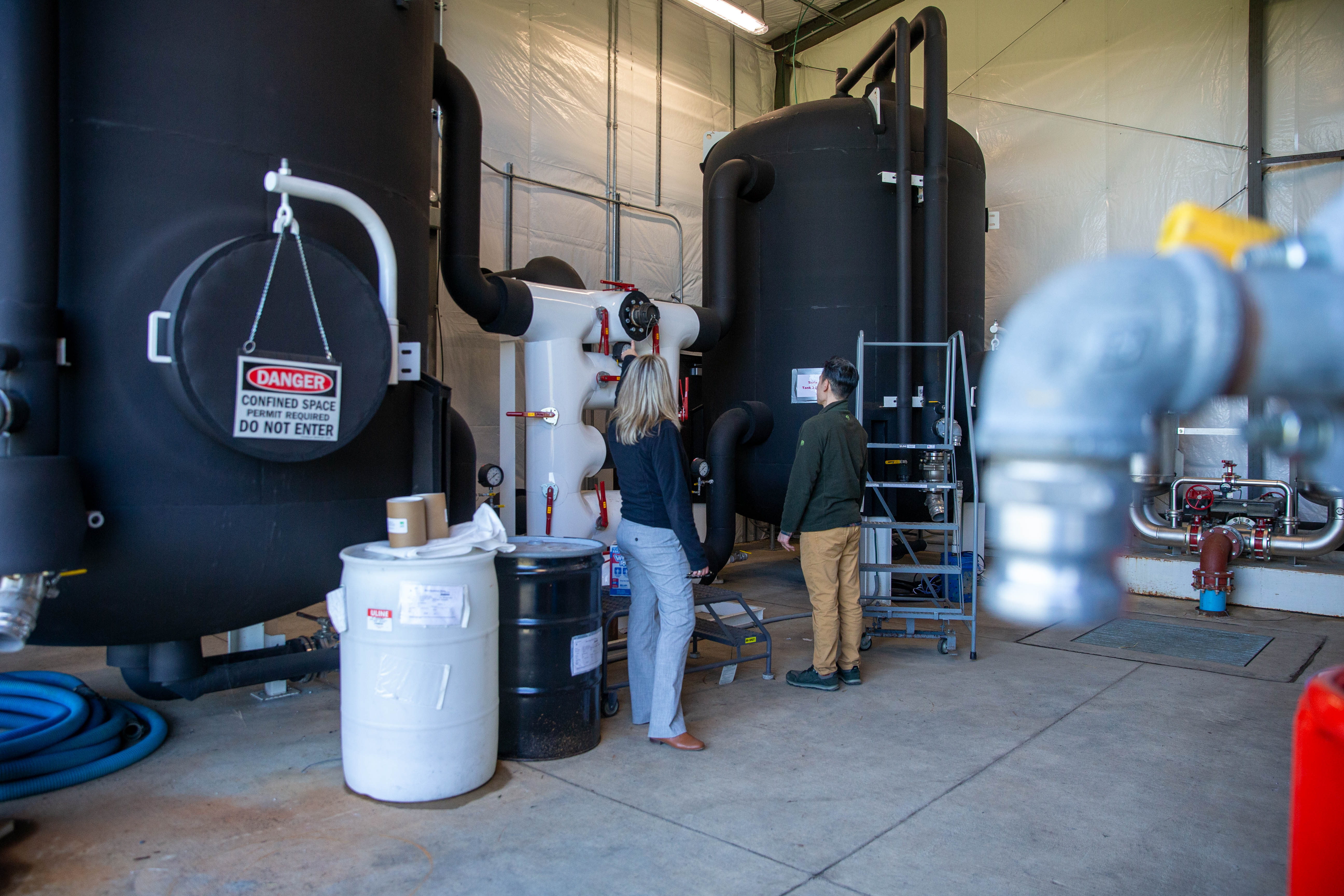 Two people look at a gauge between two large, black tanks. The tanks have two large pipes leading toward a center location. A "DANGER Confined Space" sign hangs in front of the tank.