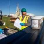A close up of soil samples in plastic containers in the foreground. Out of focus in the background is a person in a bright yellow vest and hard hat holding a notebook.